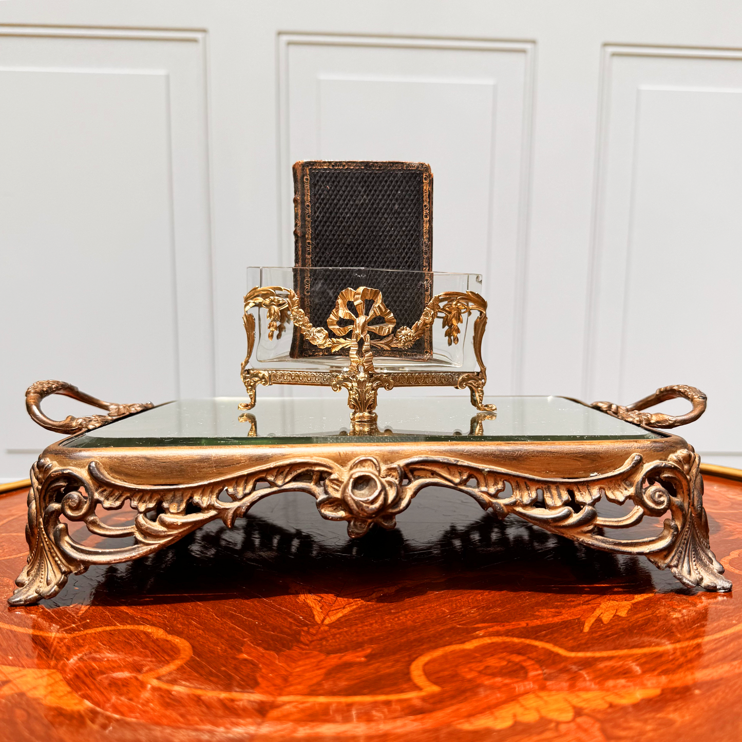 Antique mini brass-mounted crystal jardinière with diamond-shaped crystal insert, front straight-on view, placed on ornate footed mirrored riser. A small black leather book is placed inside the jardinière.