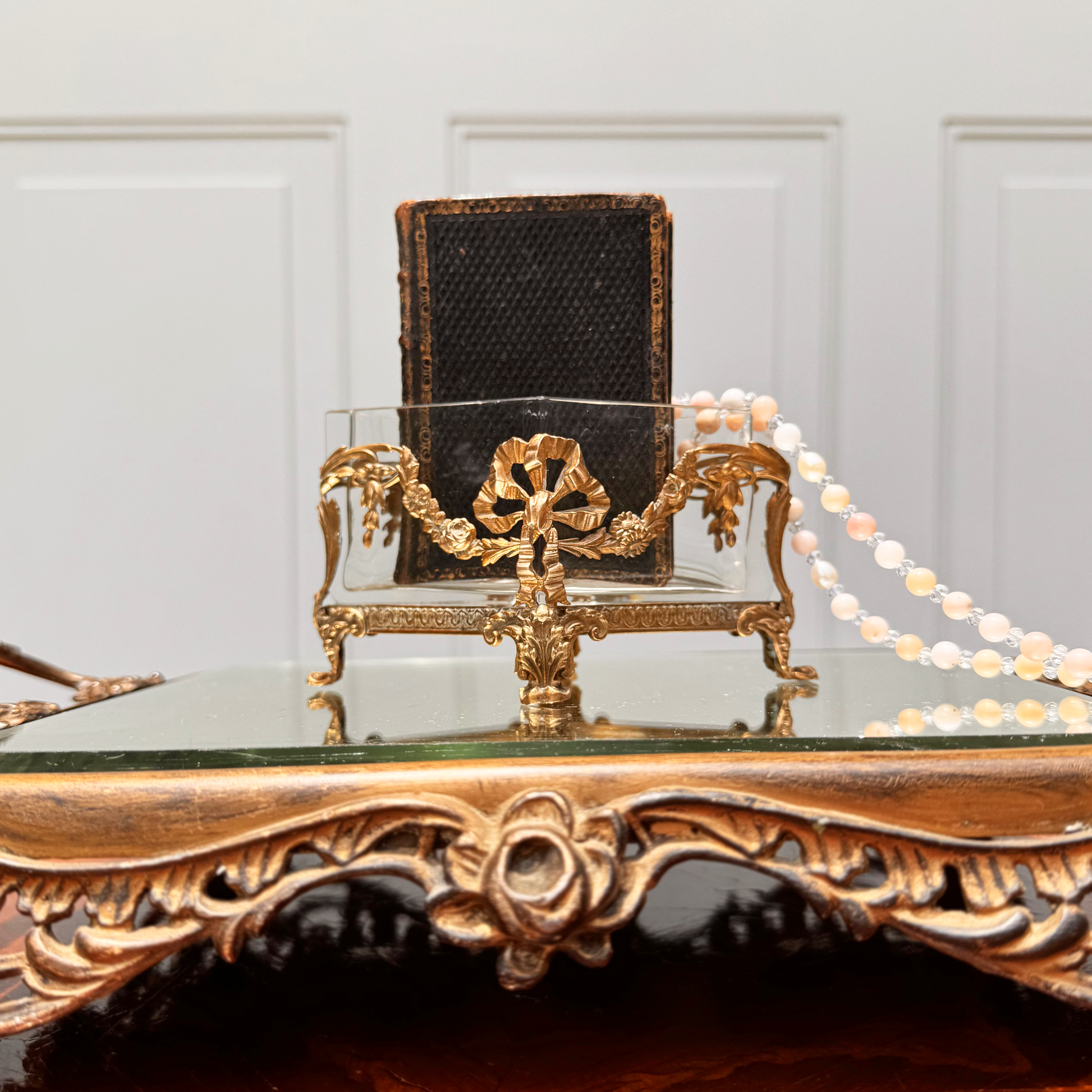Antique mini brass-mounted crystal jardinière with diamond-shaped crystal insert, front straight-on view, placed on ornate footed mirrored riser. A small black leather book is placed inside the jardinière with a pink and white beaded necklace draped over the top.