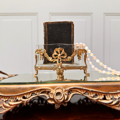 Antique mini brass-mounted crystal jardinière with diamond-shaped crystal insert, front straight-on view, placed on ornate footed mirrored riser. A small black leather book is placed inside the jardinière with a pink and white beaded necklace draped over the top.