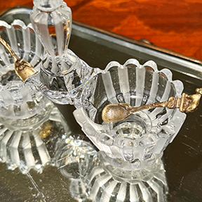 Three-quarter close-up of the Baccarat maker's mark on the inside of the crystal salt cellar, mini spoons also inside salt cellars, on mirrored surface