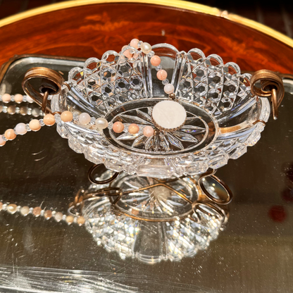Antique Baccarat crystal catchall photographed from above, showing the diamond and gemstone cut pattern, with a vintage pink, clear and white beaded necklace with a white round pendant draped inside, all placed on mirror surface