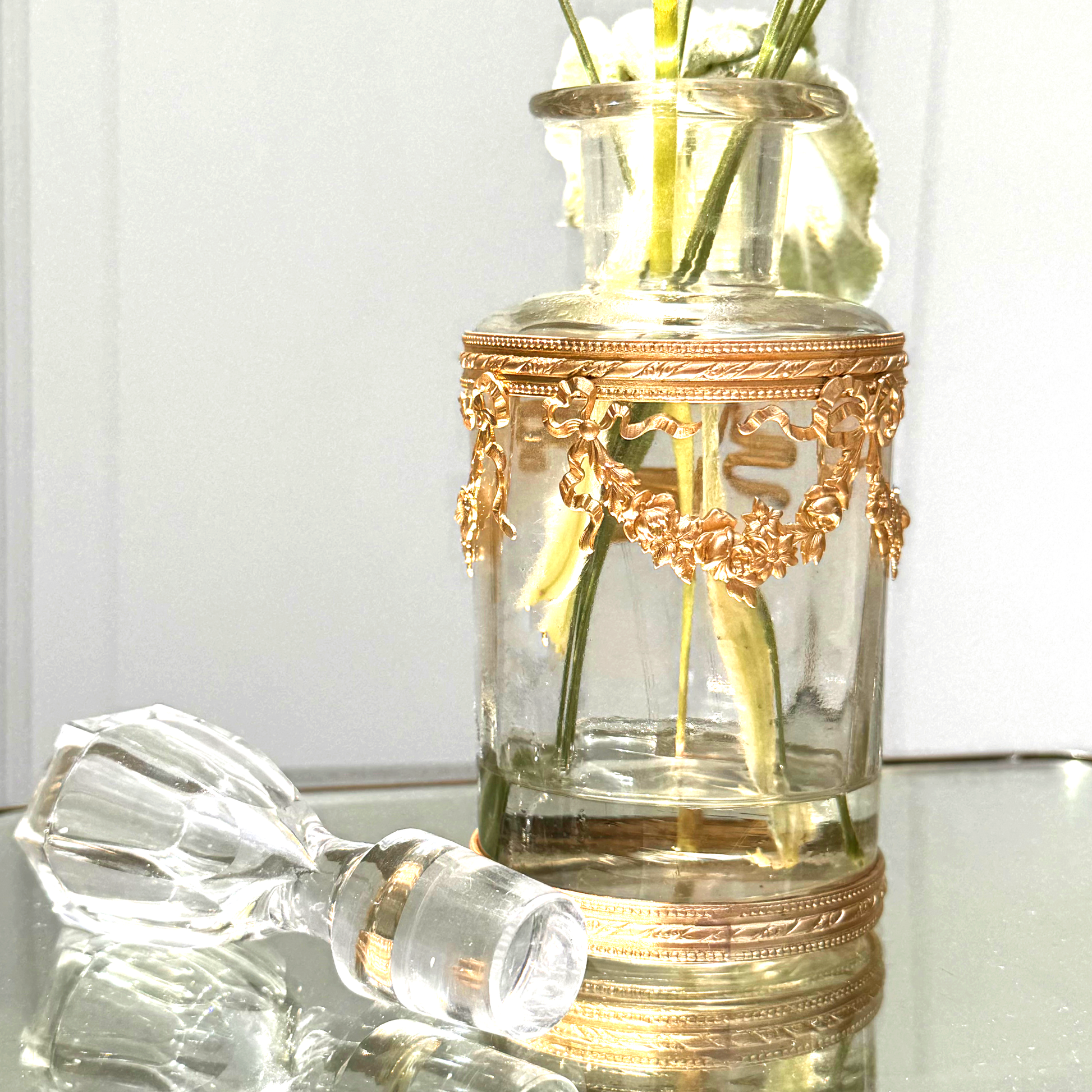 Antique ormolu-mounted blown glass vessel with crystal stopper laid beside it and fresh flowers displayed inside, angled close-up view on mirrored surface