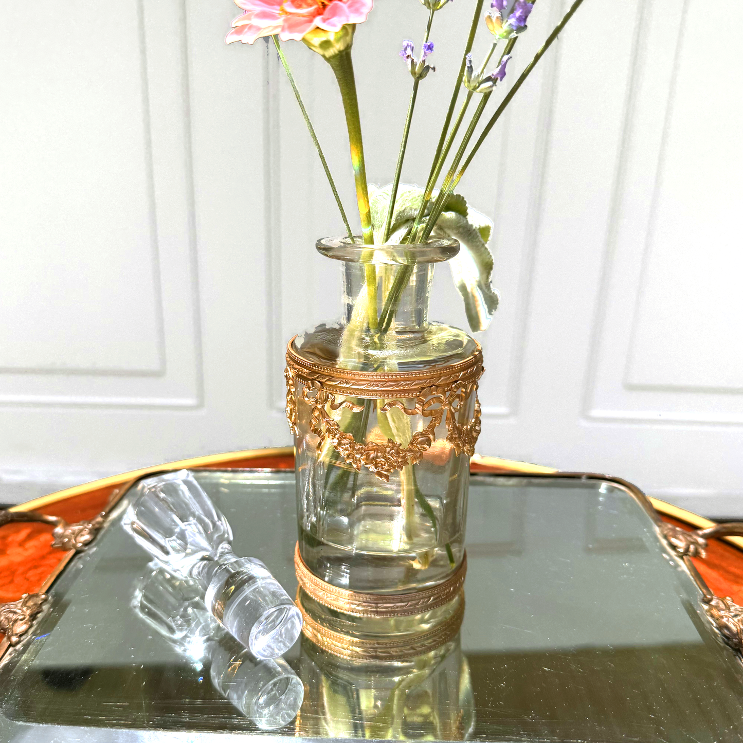 Antique ormolu-mounted blown glass vessel with crystal stopper laid beside it and fresh flowers displayed inside, front view on mirrored surface