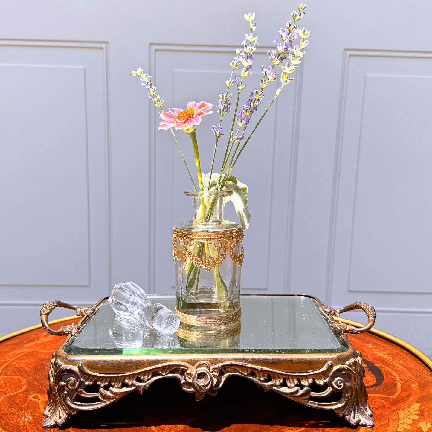 Antique ormolu-mounted blown glass vessel with crystal stopper laid beside it and fresh flowers displayed inside, front further-back view on mirrored footed tray