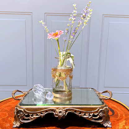 Antique ormolu-mounted blown glass vessel with crystal stopper laid beside it and fresh flowers displayed inside, front further-back view on mirrored footed tray