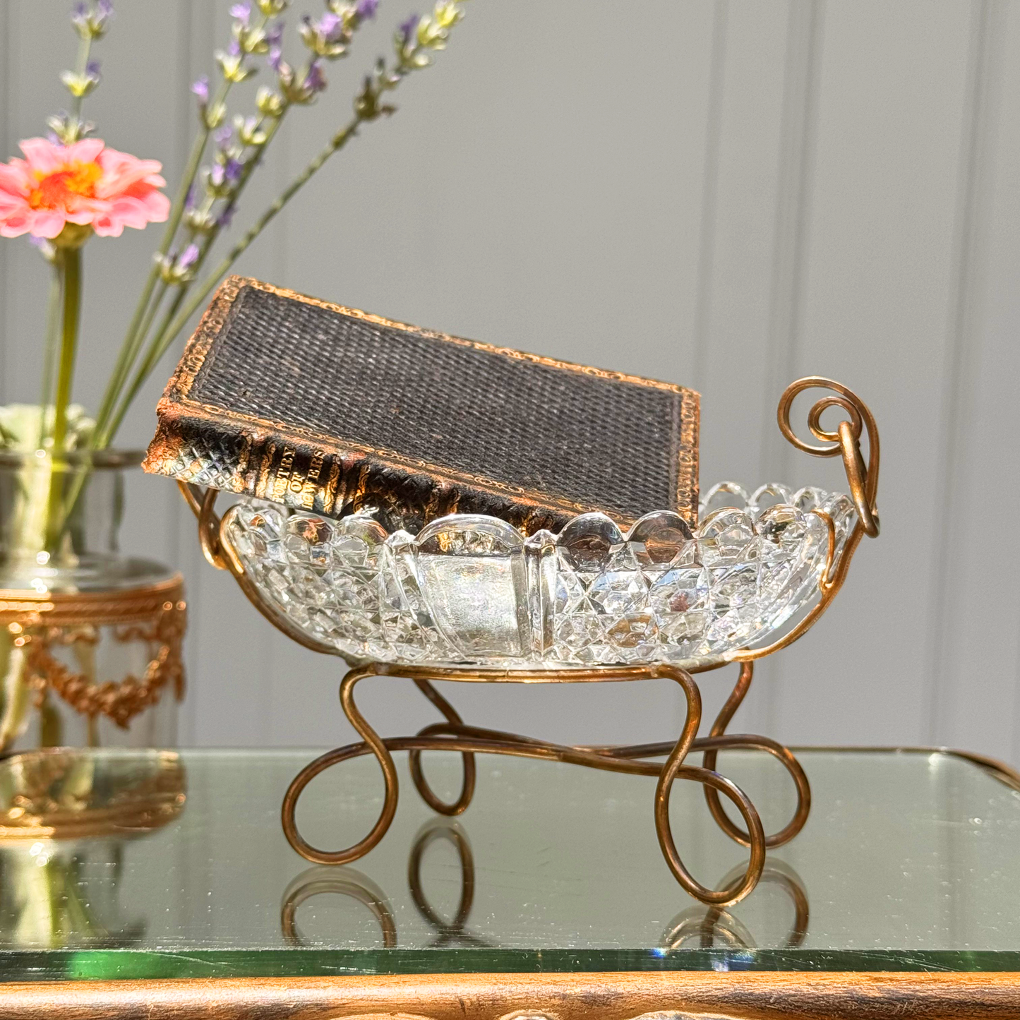 Antique Baccarat crystal catchall shown from a side angle with a miniature black and gold leather book placed inside, highlighting the curved brass mount with rings and the crystal clarity, placed on mirrored surface