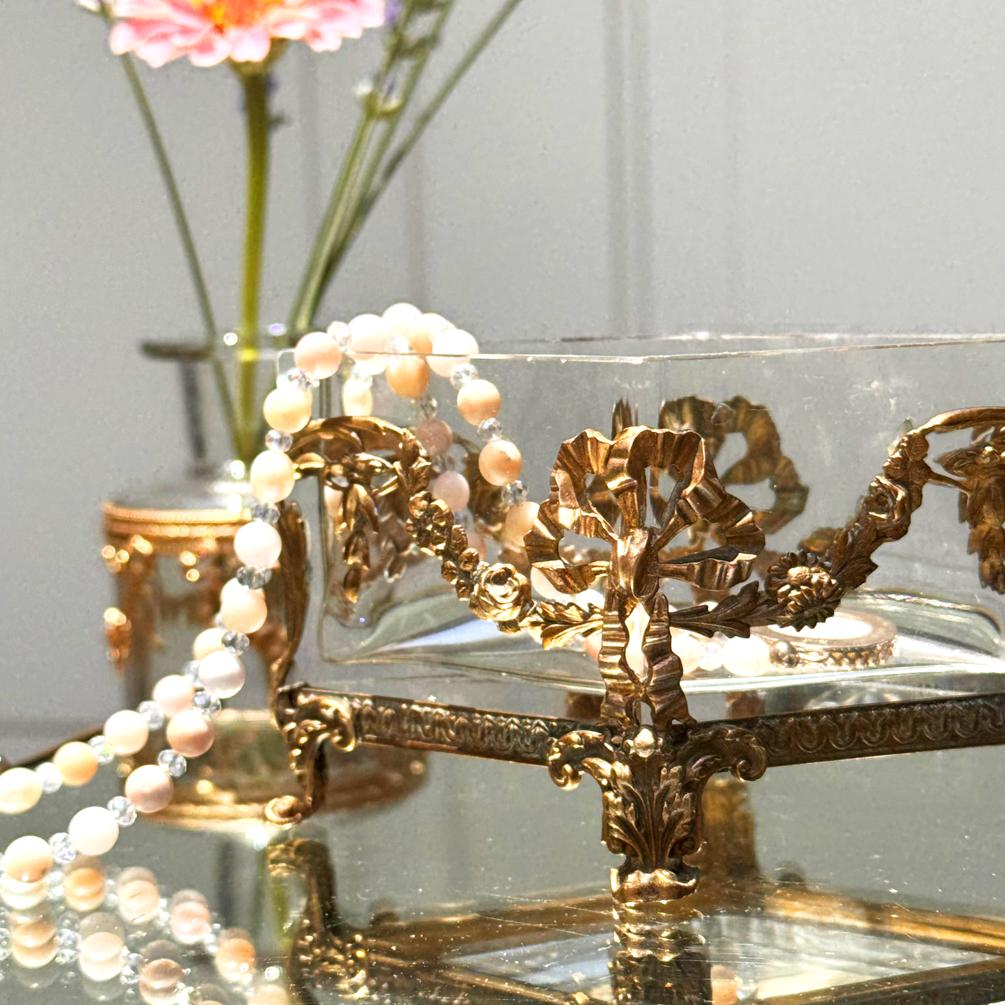 Antique brass-mounted jardinière shown in a three-quarter close-up, with a pink and white beaded necklace draped over the left side, placed upon a mirrored surface