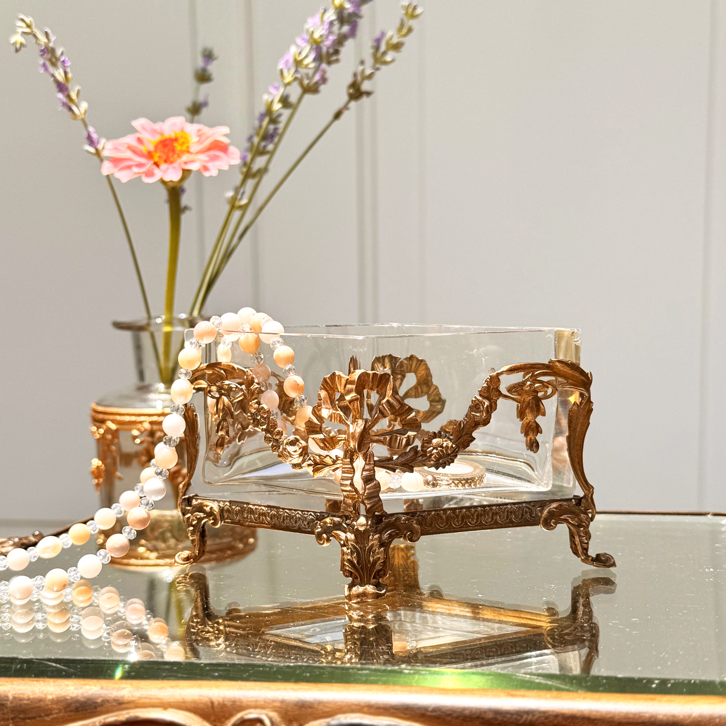 Antique brass-mounted jardinière shown straight on, with a pink and white beaded necklace draped over the left side, placed upon a mirrored surface