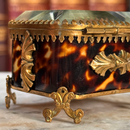 An antique miniature footed tortoiseshell jewelry casket with brass mounts and a beveled glass top, resting on a marble surface, shown closed from side/back angle highlighting foot and shell