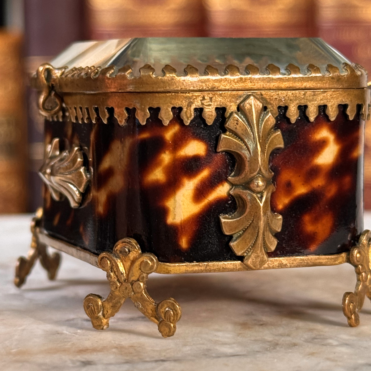 An antique miniature footed tortoiseshell jewelry casket with brass mounts and a beveled glass top, resting on a marble surface, shown closed from side highlighting shell and adornments
