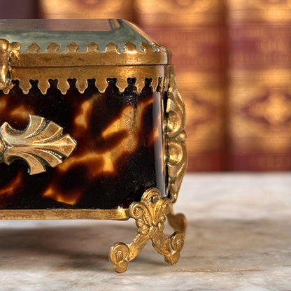 An antique miniature footed tortoiseshell jewelry casket with brass mounts and a beveled glass top, resting on a marble surface, shown closed from front highlighting one half of the box