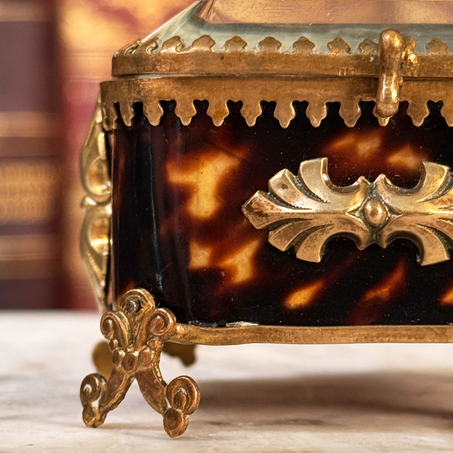 An antique miniature footed tortoiseshell jewelry casket with brass mounts and a beveled glass top, resting on a marble surface, shown closed from front highlighting shell and adornments on one half