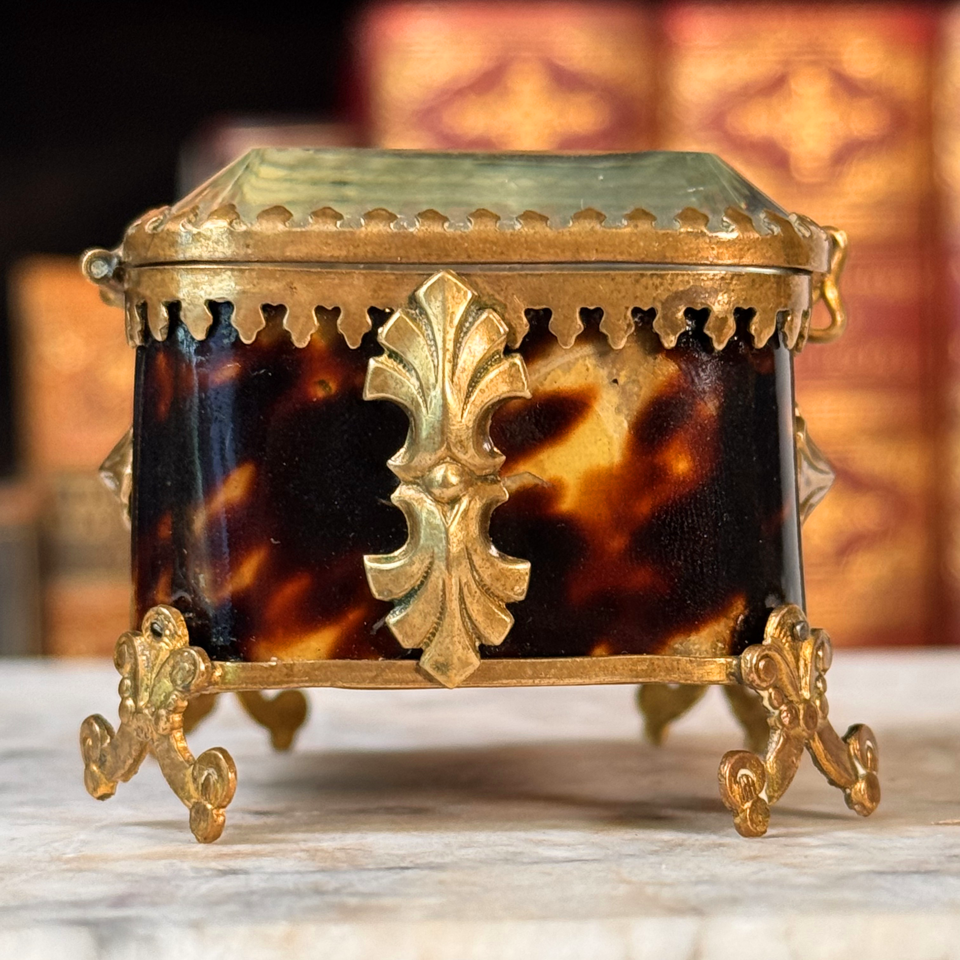 An antique miniature footed tortoiseshell jewelry casket with brass mounts and a beveled glass top, resting on a marble surface, shown closed from side highlighting shell and adornments, close-up