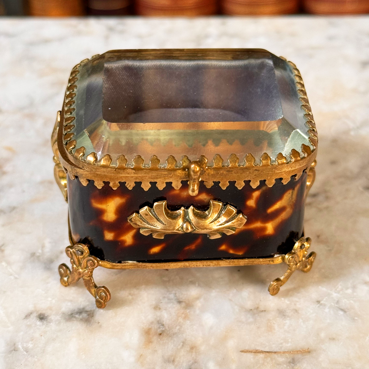 An antique miniature footed tortoiseshell jewelry casket with brass mounts and a beveled glass top, resting on a marble surface, angled overhead view highlighted beveled glass lid