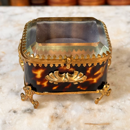 An antique miniature footed tortoiseshell jewelry casket with brass mounts and a beveled glass top, resting on a marble surface, angled overhead view highlighted beveled glass lid