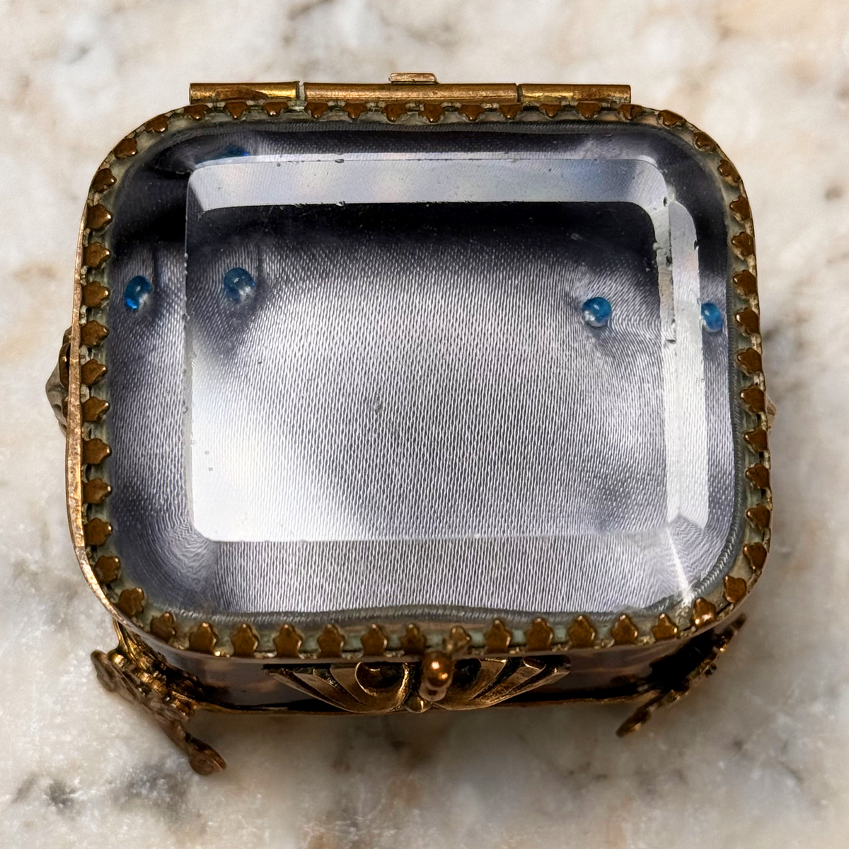 An antique miniature footed tortoiseshell jewelry casket with brass mounts and a beveled glass top, resting on a marble surface, overhead view close-up highlighting interior and beveled glass lid