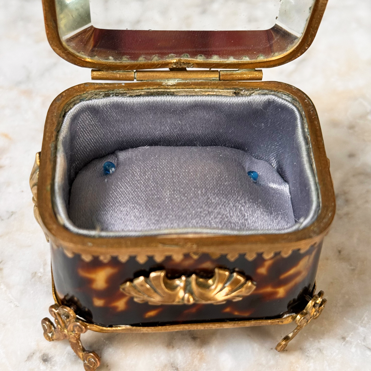 An antique miniature footed tortoiseshell jewelry casket with brass mounts and a beveled glass top, resting on a marble surface, shown open from front highlighting interior