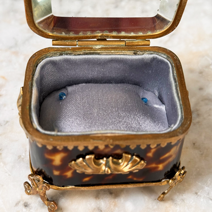 An antique miniature footed tortoiseshell jewelry casket with brass mounts and a beveled glass top, resting on a marble surface, shown open from front highlighting interior