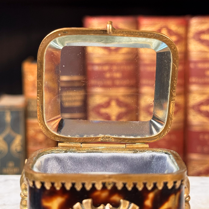 An antique miniature footed tortoiseshell jewelry casket with brass mounts and a beveled glass top, resting on a marble surface, shown open from front highlighting interior of beveled glass lid