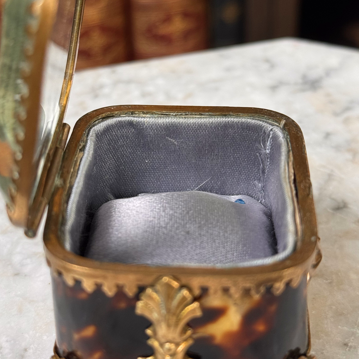 An antique miniature footed tortoiseshell jewelry casket with brass mounts and a beveled glass top, resting on a marble surface, shown open from side highlighted interior