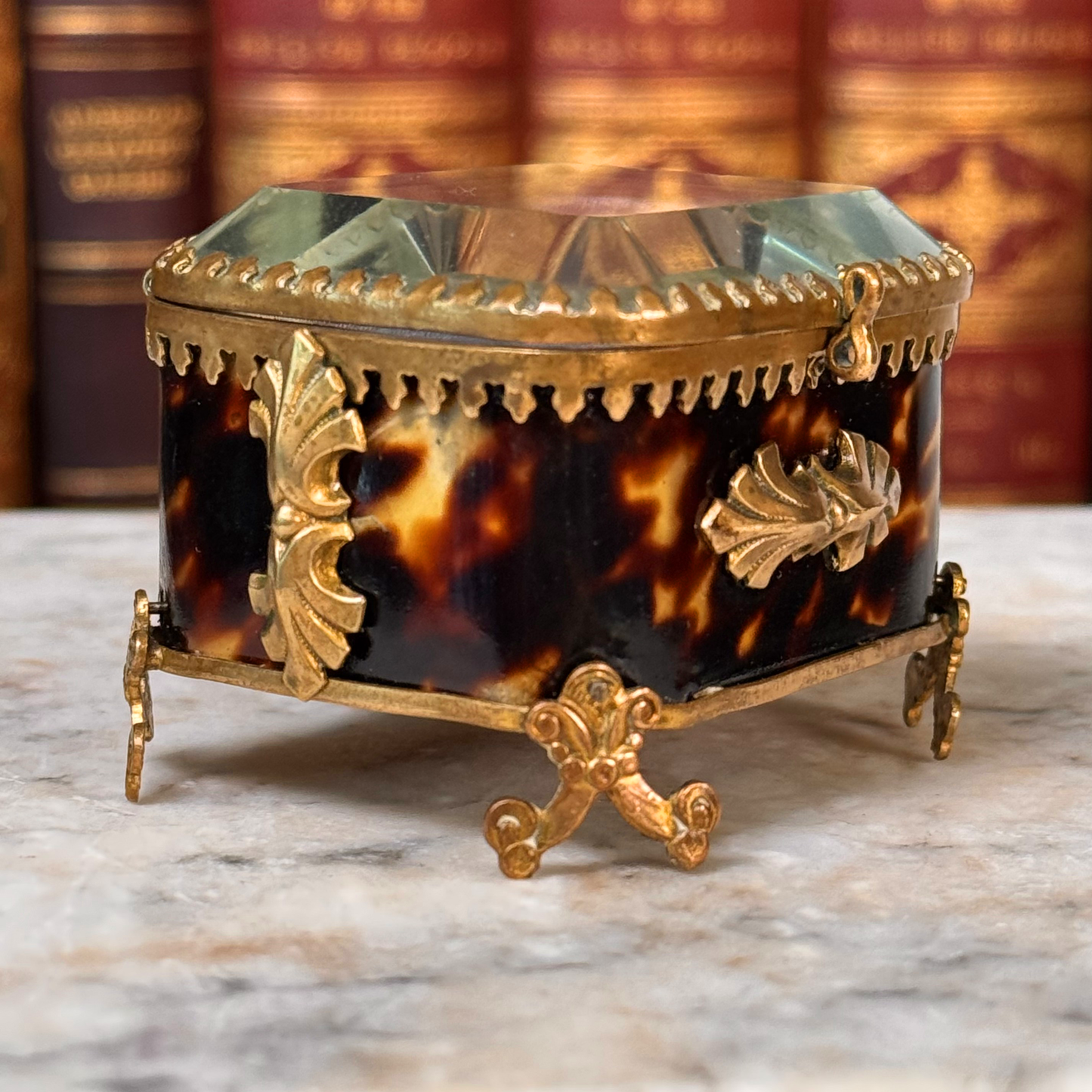An antique miniature footed tortoiseshell jewelry casket with brass mounts and a beveled glass top, resting on a marble surface, shown closed from angled side highlighting shell and adornments