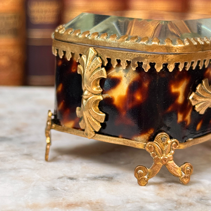 An antique miniature footed tortoiseshell jewelry casket with brass mounts and a beveled glass top, resting on a marble surface, shown closed from angled side highlighting shell and adornments close-up