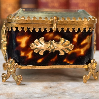 An antique miniature footed tortoiseshell jewelry casket with brass mounts and a beveled glass top, resting on a marble surface, shown closed from side highlighting shell and adornments shown close-up from front straight on