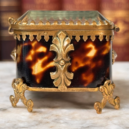 An antique miniature footed tortoiseshell jewelry casket with brass mounts and a beveled glass top, resting on a marble surface, shown closed from side highlighting shell and adornments, close-up