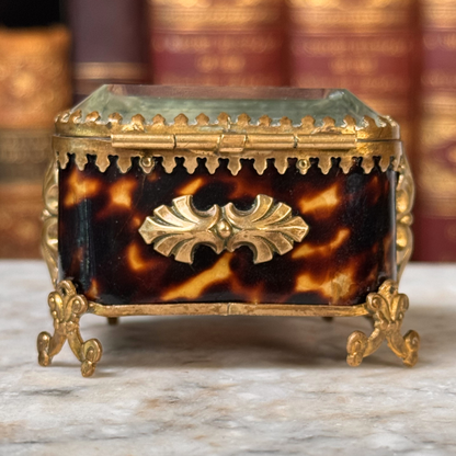 An antique miniature footed tortoiseshell jewelry casket with brass mounts and a beveled glass top, resting on a marble surface, straight on close-up view of backside