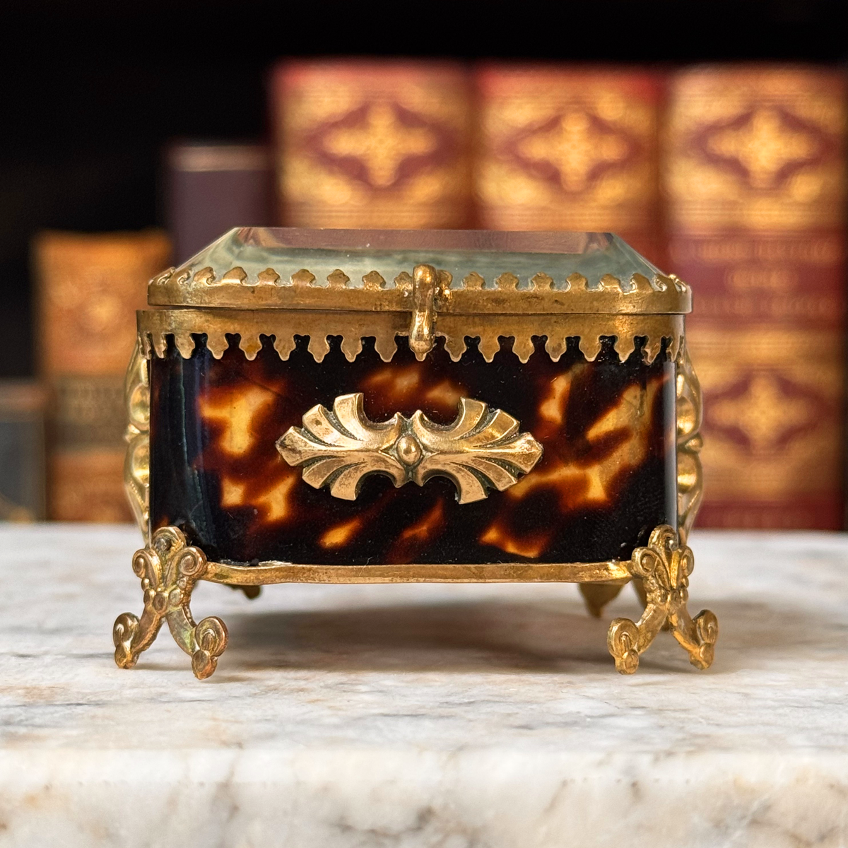 An antique miniature footed tortoiseshell jewelry casket with brass mounts and a beveled glass top, resting on a marble surface, straight on close-up view