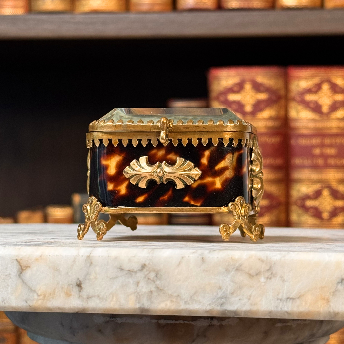An antique miniature footed tortoiseshell jewelry casket with brass mounts and a beveled glass top, resting on a marble surface. Slightly angled from below view.