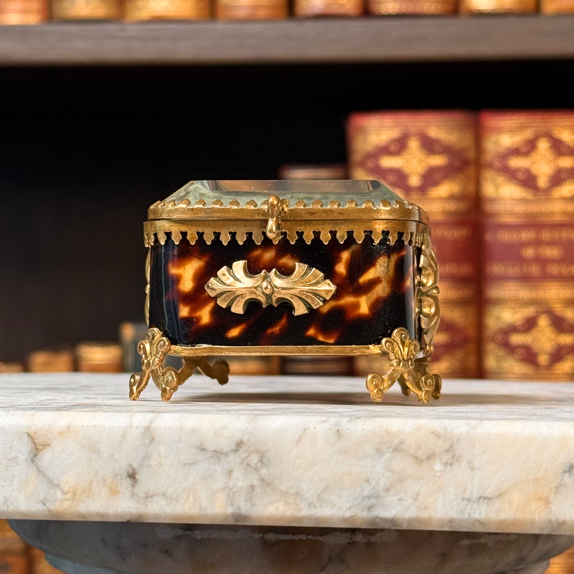An antique miniature footed tortoiseshell jewelry casket with brass mounts and a beveled glass top, resting on a marble surface. Slightly angled from below view.