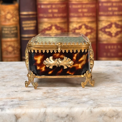 An antique miniature footed tortoiseshell jewelry casket with brass mounts and a beveled glass top, resting on a marble surface. Straight on view