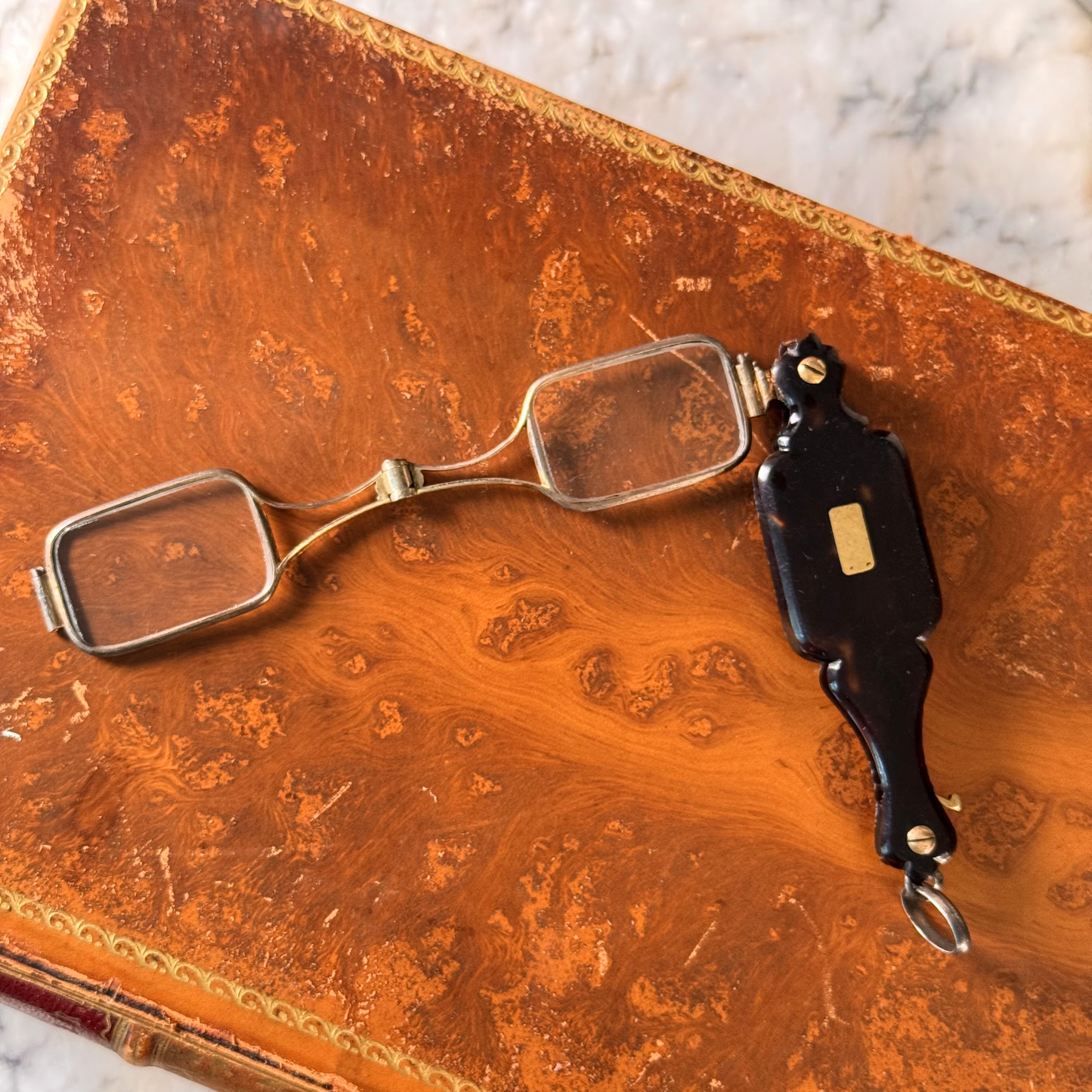 An antique folding lorgnette and spectacles shown unfolded with a tortoiseshell handle and silver metal frames, resting on an antique brown leather book cover