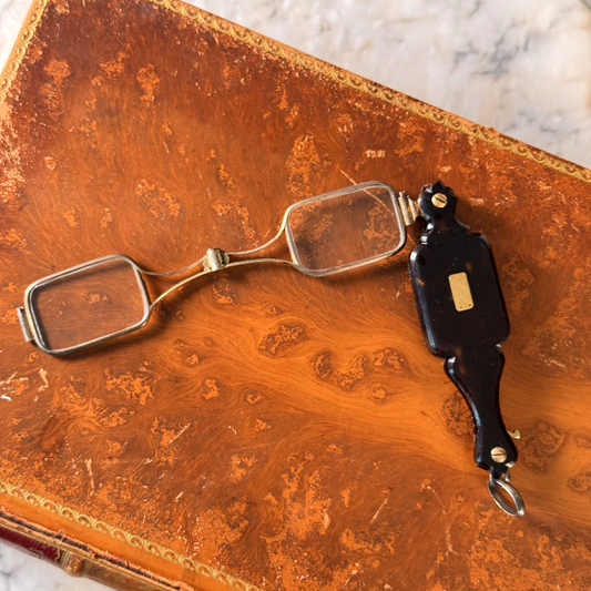 An antique folding lorgnette and spectacles shown unfolded with a tortoiseshell handle and silver metal frames, resting on an antique brown leather book cover