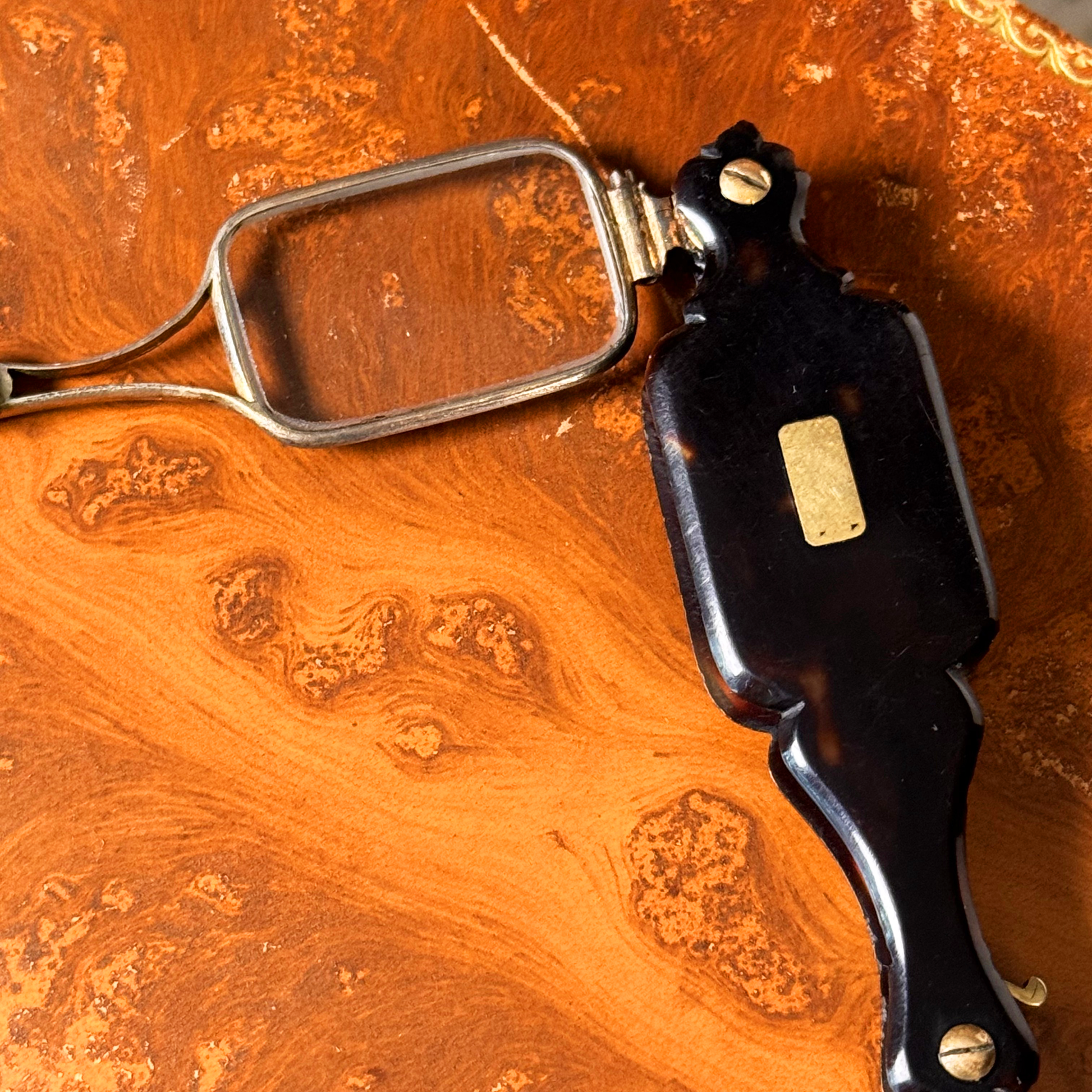 An antique folding lorgnette and spectacles shown unfolded with a tortoiseshell handle, silver colored frames, and gold metal plaque, resting on a leather book, close-up on one side of frames and handle