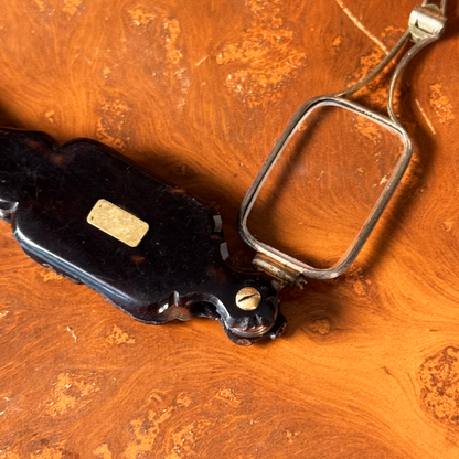 An antique folding lorgnette and spectacles shown unfolded with a tortoiseshell handle, silver colored frames,  and gold plaque, resting on a leather book, close-up of front of handle