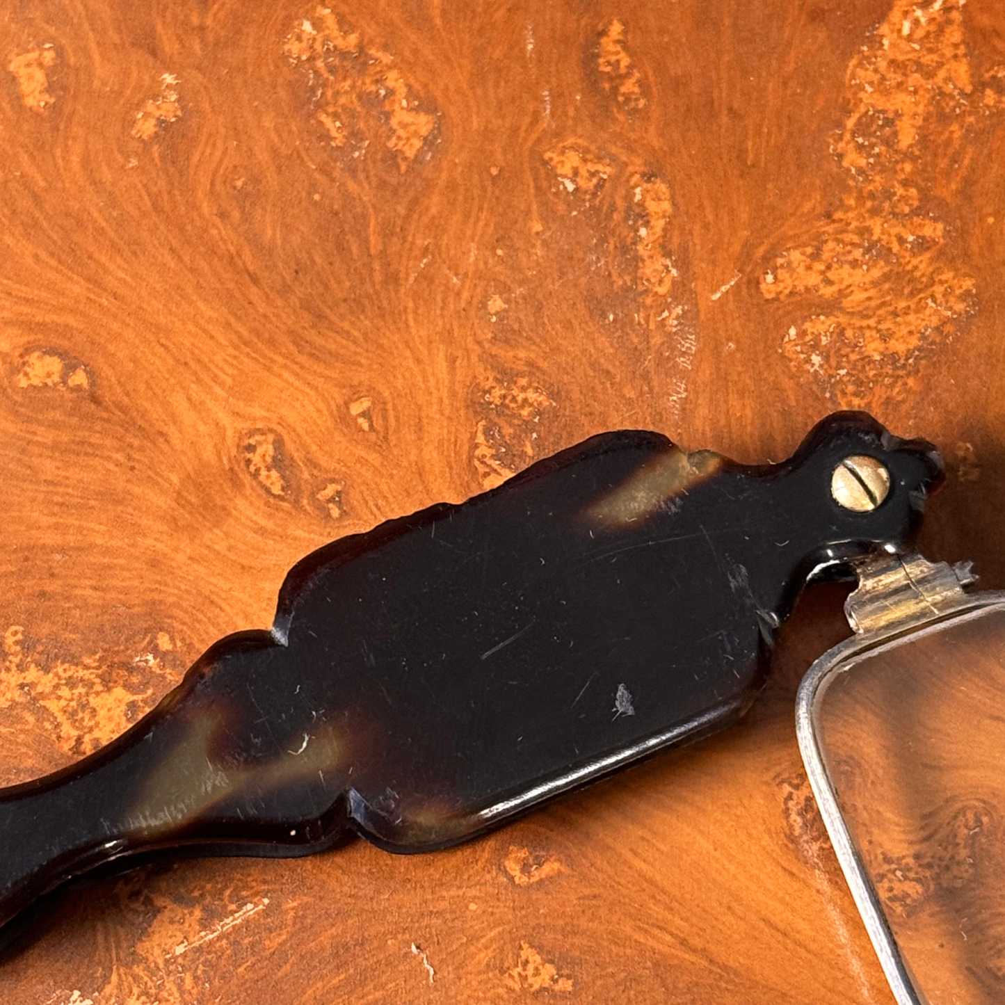 An antique folding lorgnette and spectacles shown unfolded with a tortoiseshell handle, silver colored frames,  resting on a leather book, close-up of backside of handle, showing chipping to edges