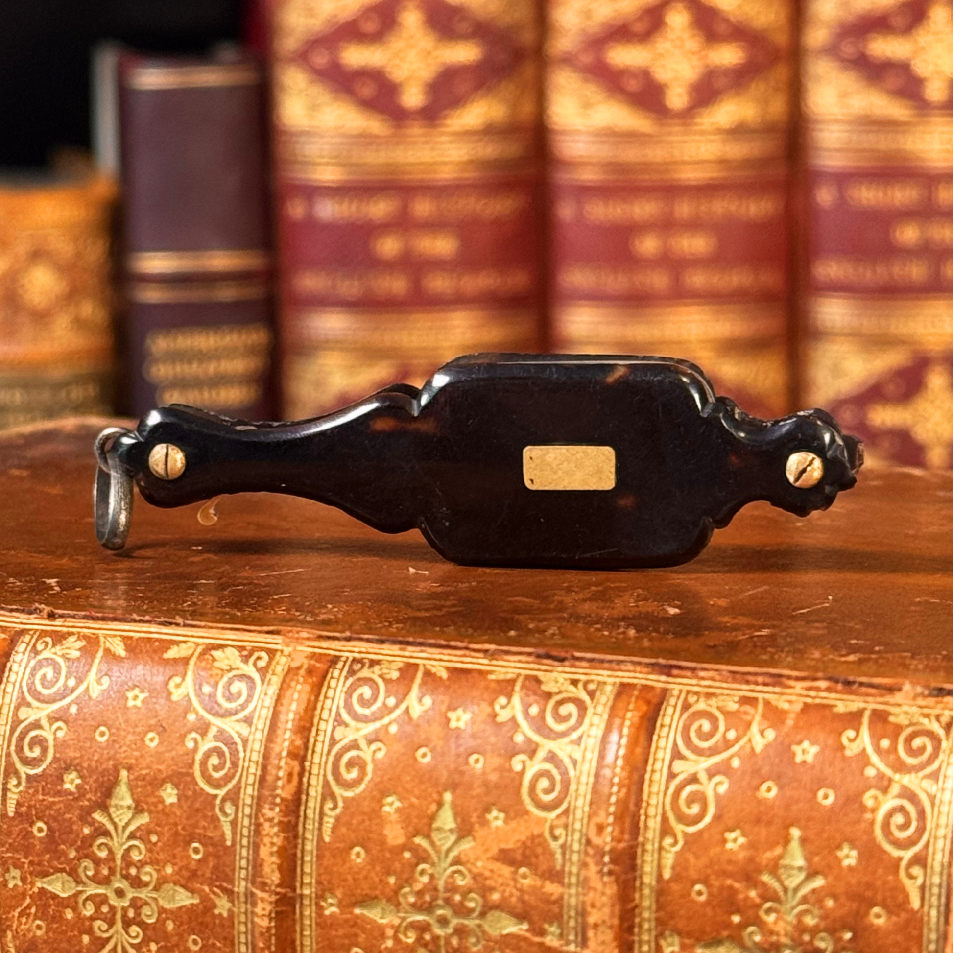 An antique folding lorgnette and spectacles shown folded with a tortoiseshell handle, gold plaque, and silver colored frames, resting on a leather book, front shown