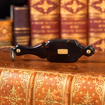 An antique folding lorgnette and spectacles shown folded with a tortoiseshell handle, gold plaque, and silver colored frames, resting on a leather book, front shown