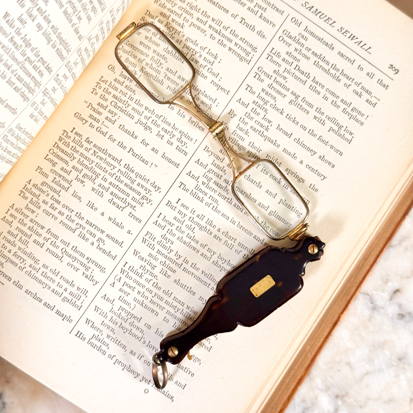 An antique folding lorgnette and spectacles shown unfolded with a tortoiseshell handle, silver colored frames, and gold metal plaque, resting on the inside of an open book