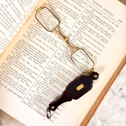 An antique folding lorgnette and spectacles shown unfolded with a tortoiseshell handle, silver colored frames, and gold metal plaque, resting on the inside of an open book