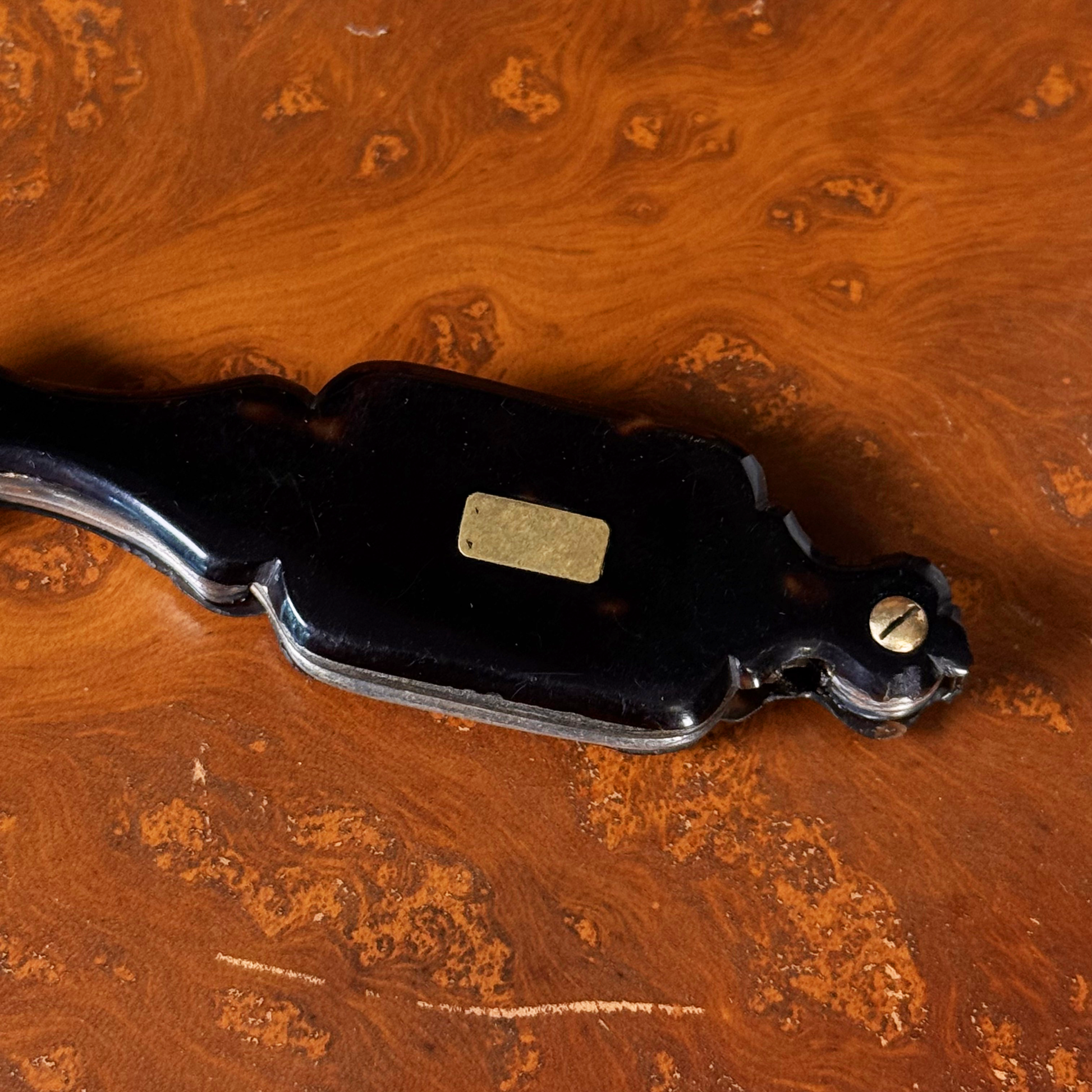 An antique folding lorgnette and spectacles shown folded with a tortoiseshell handle, gold plaque, and silver colored frames, resting on a leather book, close up on plaque and top front