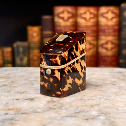 An antique tortoiseshell box with a glossy dark brown and amber pattern, featuring a gold-toned metal piece on top and a small round button, shown from side angle