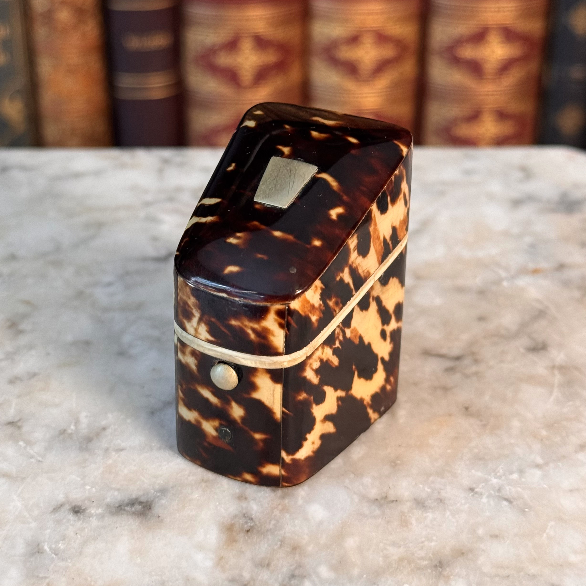 An antique tortoiseshell box with a glossy dark brown and amber pattern, featuring a gold-toned metal piece on top and a small round button