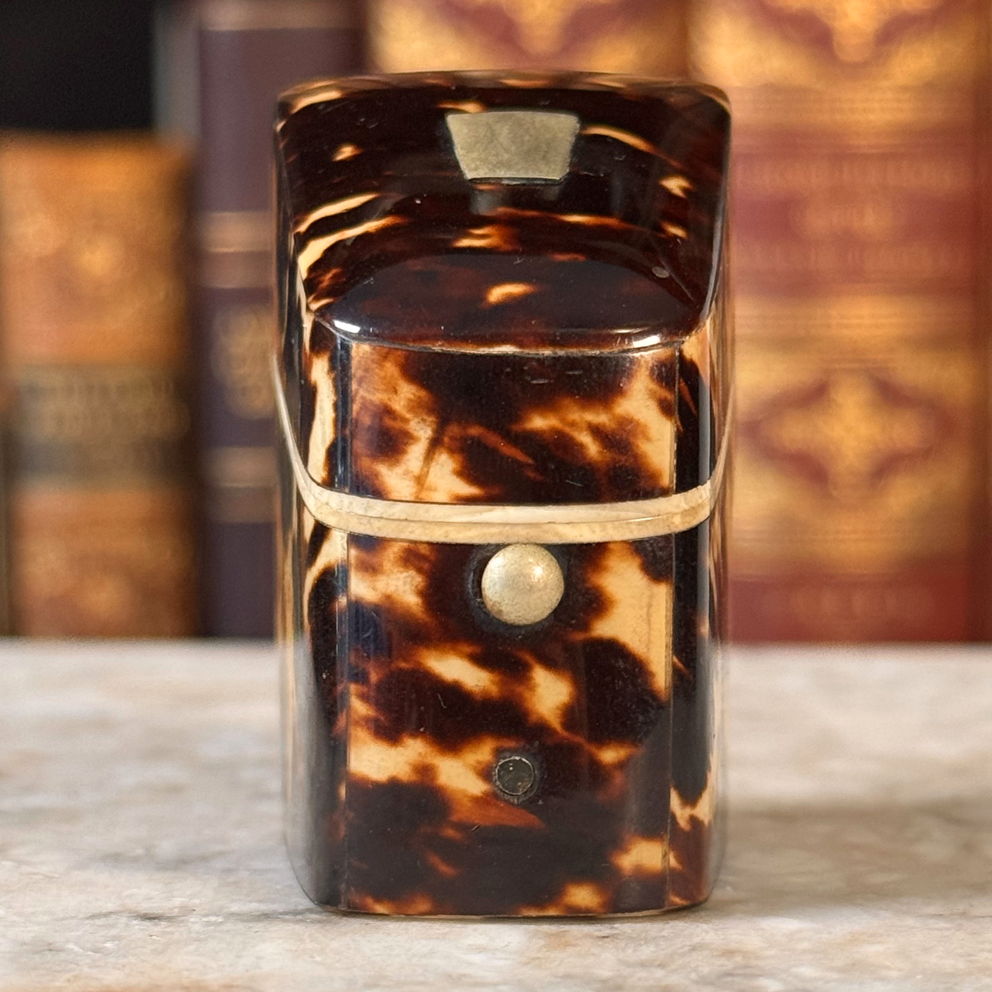 An antique tortoiseshell box with a glossy dark brown and amber pattern, featuring a gold-toned metal piece on top and a small round button, shown from front straight on