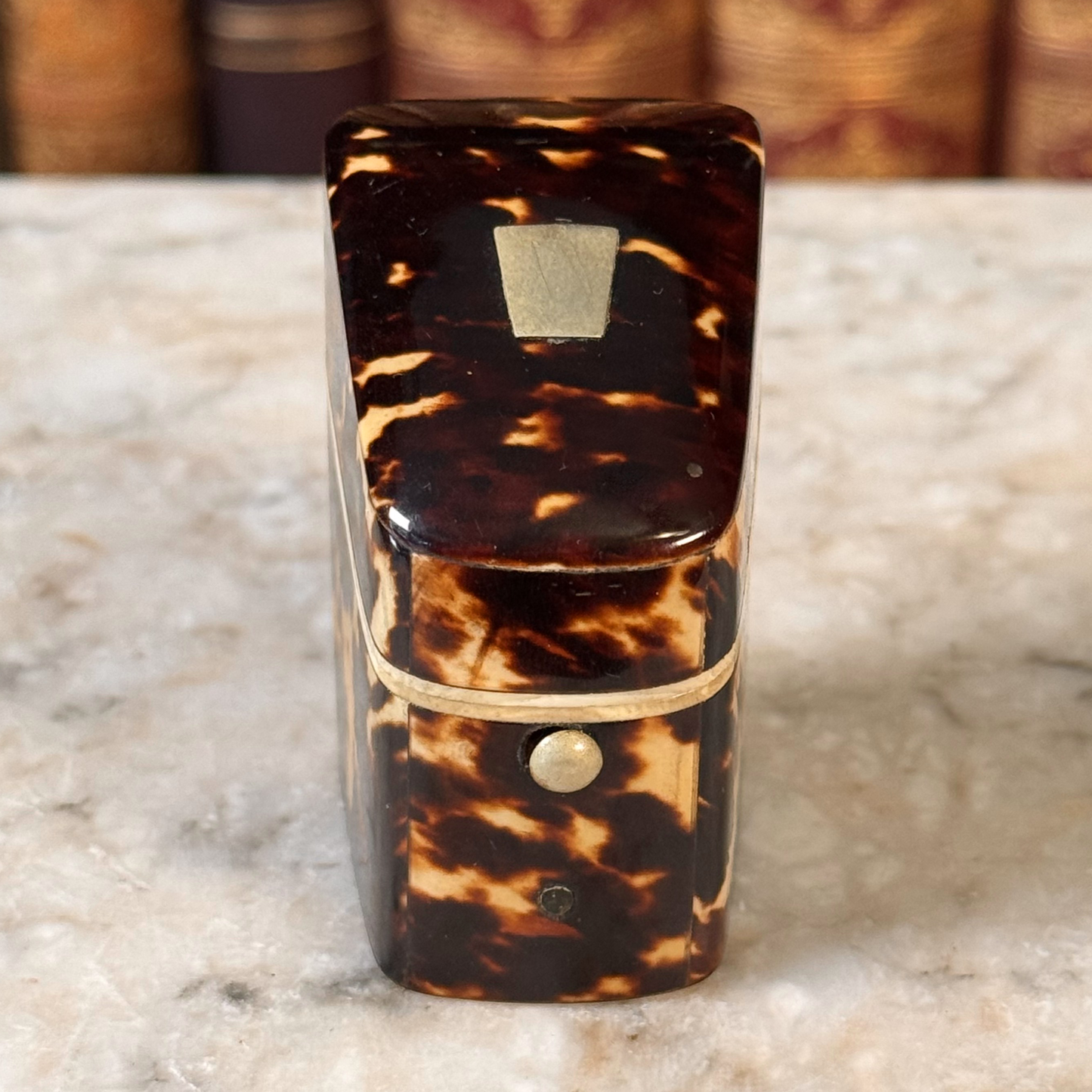 An antique tortoiseshell box with a glossy dark brown and amber pattern, featuring a gold-toned metal piece on top and a small round button, shown from front angled above