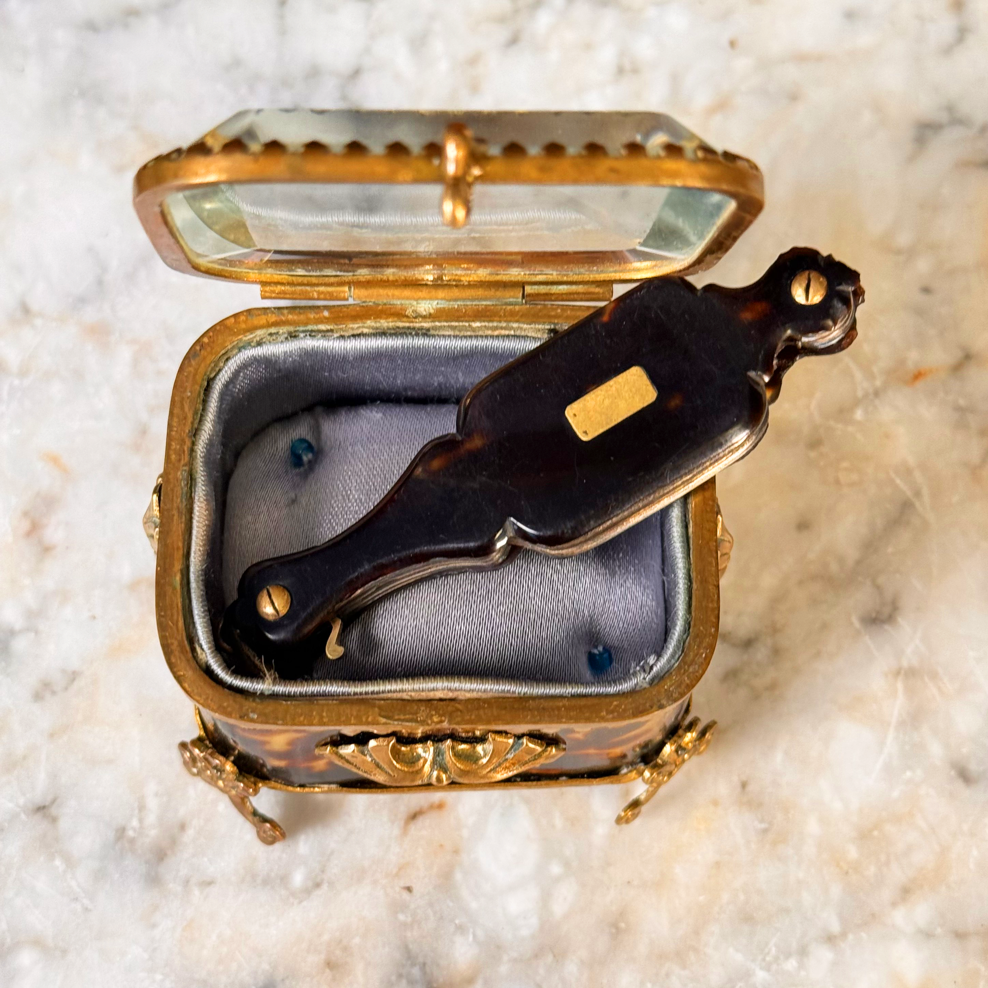 An antique folding lorgnette and spectacles shown folded with a tortoiseshell handle, gold plaque, and silver colored frames, placed inside a mini jewelry box