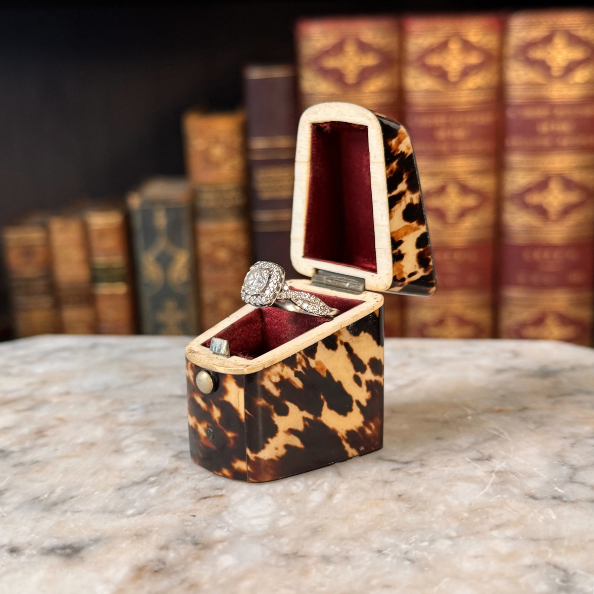 An antique tortoiseshell box with a glossy dark brown and amber pattern, a small round button, shown open with red silk lining on top and bottom and a ring displayed inside