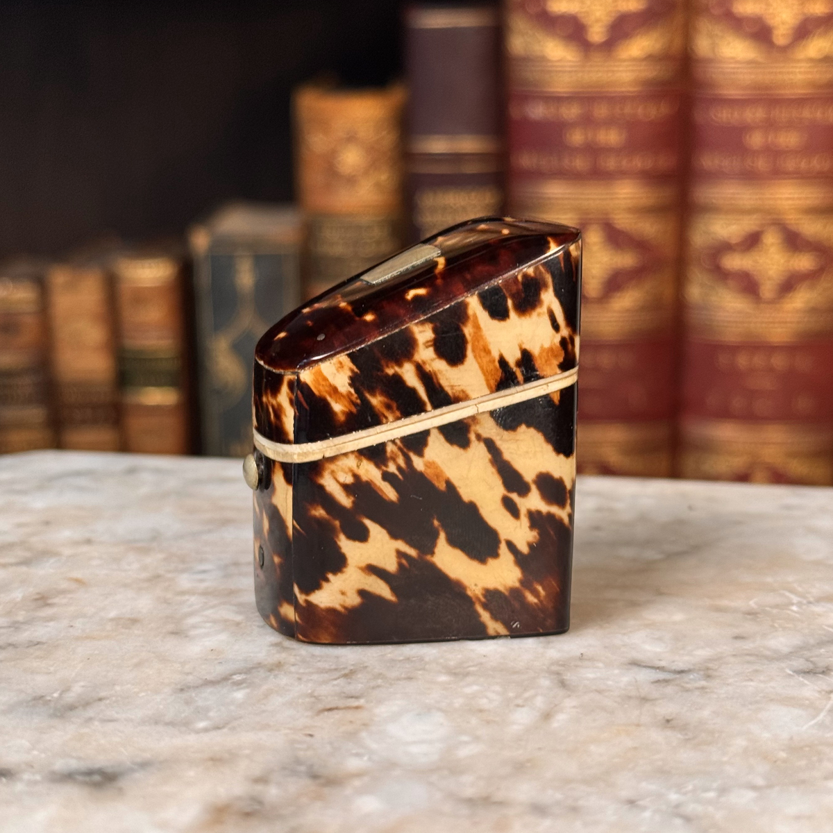 An antique tortoiseshell box with a glossy dark brown and amber pattern, featuring a gold-toned metal piece on top and a small round button, shown from one side